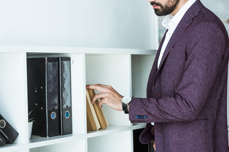 cropped shot of businessman taking book from shelf at officeの写真素材