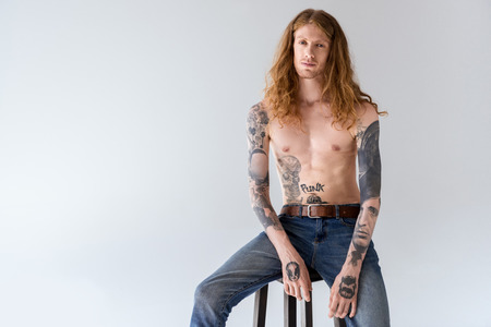 handsome shirtless tattooed man with curly hair sitting on chair and looking at camera isolated on whiteの写真素材
