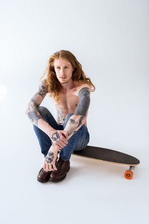 handsome shirtless tattooed skater sitting on longboard with curly hairの写真素材