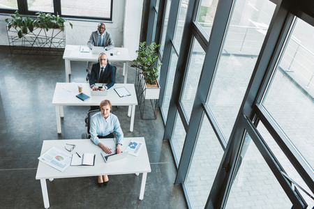 high angle view of multiethnic business people sitting at tables and looking at camera in modern...