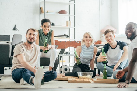 smiling multiethnic friends sitting on floor with bottles of beer, pizza and popcornの写真素材