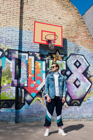 handsome young man in vintage clothing in front of brick wall with graffiti and basketball ringの写真素材