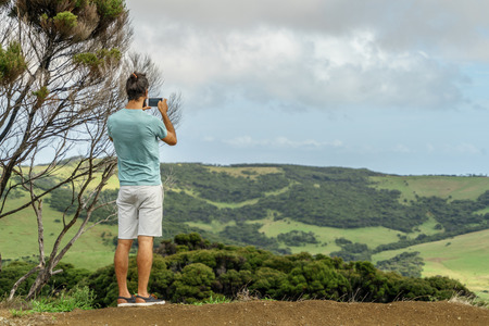 rear view of man taking photo of beautiful green valley with smartphone, New Zealandの写真素材