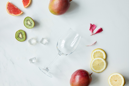 flat lay with empty wineglass, flower petals, ice cubes and assorted exotic fruits on white tabletopの写真素材