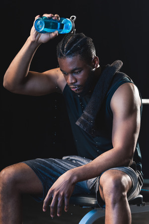 tired african american sportsman pouring water on head while sitting after workout in gymの写真素材