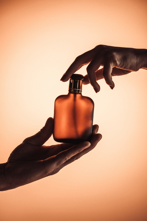 cropped view of man and woman holding one bottle of perfume, isolated on orangeの写真素材