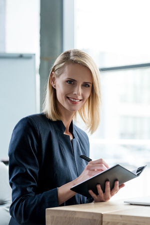 beautiful businesswoman smiling at camera while taking notes in notebookの写真素材