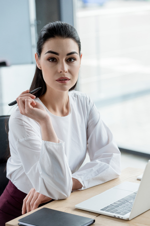 beautiful young businesswoman holding pen and looking at camera in officeの写真素材
