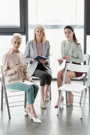 beautiful businesswomen sitting on chairs and looking at camera in officeの写真素材
