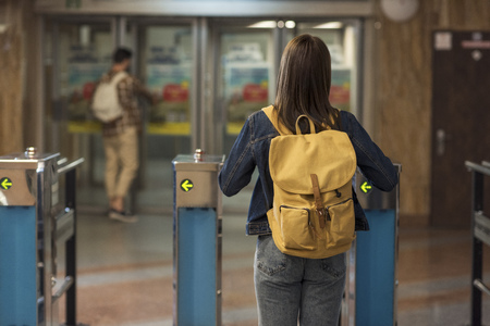 rear view of female stylish traveler with backpack passing through turnstiles and male tourist...
