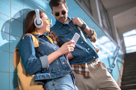 smiling stylish female tourist with headphones showing smartphone to boyfriend in sunglassesの写真素材