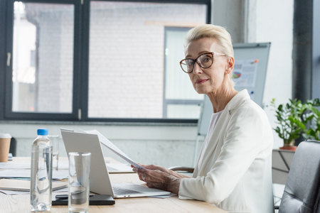 attractive senior businesswoman in eyeglasses holding documents and looking at camera in officeの写真素材