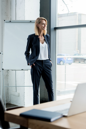 stylish blonde businesswoman standing with hands in pockets and looking at windowの写真素材