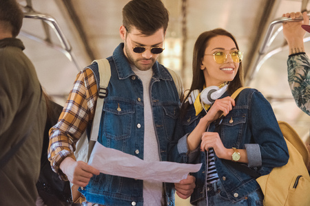 young stylish female traveler in sunglasses with headphones and boyfriend looking at map in metro trainの写真素材