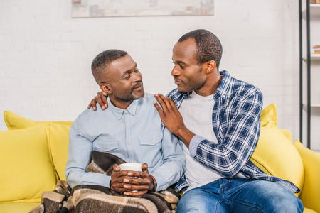 smiling senior man with cup of hot beverage sitting under plaid and looking at adult son at home