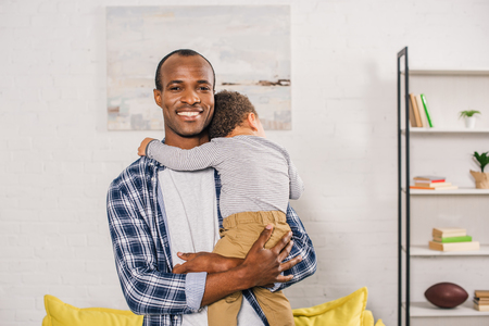 happy young african american father smiling at camera while carrying adorable little son at homeの写真素材