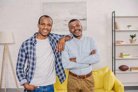 happy african american father and adult son standing together and smiling at camera at home