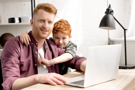 happy father and son smiling at camera while using laptop together at homeの写真素材