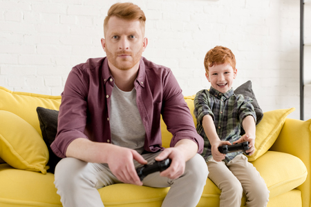 father and son sitting on couch and playing with joysticks at homeの写真素材