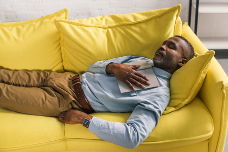 high angle view of senior african american man sleeping with book on sofaの写真素材