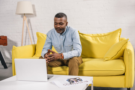 senior african american man sitting on sofa and looking at laptopの写真素材