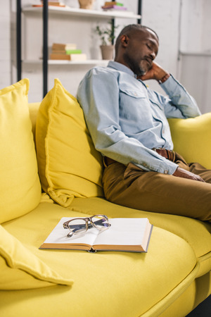 book and eyeglasses on sofa and senior man sleeping behindの写真素材