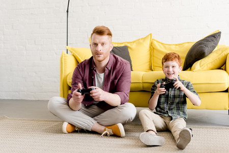 happy father and son sitting on carpet and playing with joysticks at home