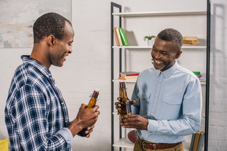 smiling african american father with adult son holding beer bottles at homeの写真素材