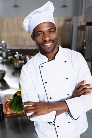 Smiling african american chef standing with arms folded in modern kitchenの写真素材