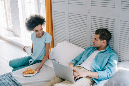 African american woman having breakfast by her boyfriend in bed with laptopの写真素材