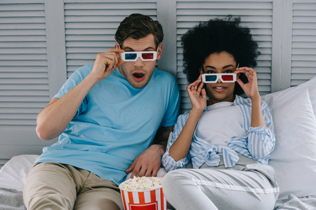 Multiracial boyfriend and girlfriend in stereoscopic glasses watching movie at home with popcornの写真素材