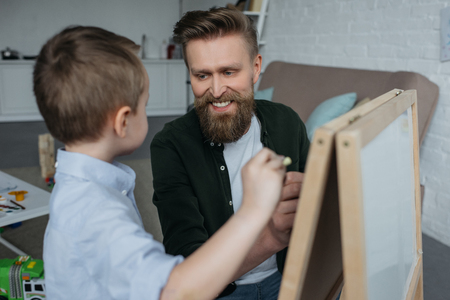 little boy and smiling father with pieces of chalk drawing picture on blackboard at homeの写真素材