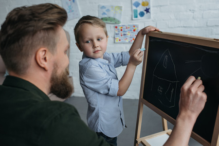 side view of little boy and father with pieces of chalk drawing picture on blackboard at homeの写真素材
