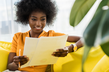 portrait of smiling african american woman with papers remote working on sofa at homeの写真素材