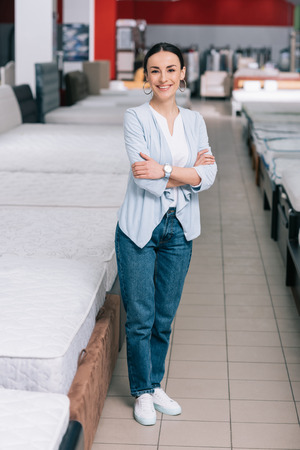 smiling female customer with arms crossed standing in furniture store with arranged mattressesの写真素材
