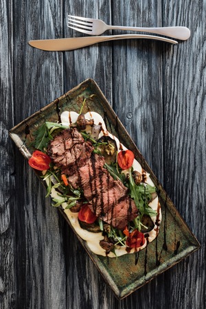 top view of delicious roasted sliced New York steak, vegetables and fork with knife on wooden tableの写真素材