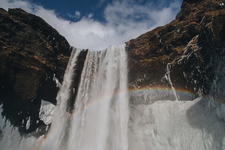 low angle view of beautiful skogafoss waterfall with frozen icicles on rocks and rainbow in icelandの写真素材
