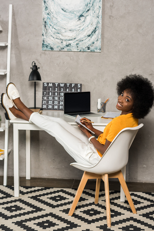 smiling african american woman with notebook sitting at table with laptop at home officeの写真素材