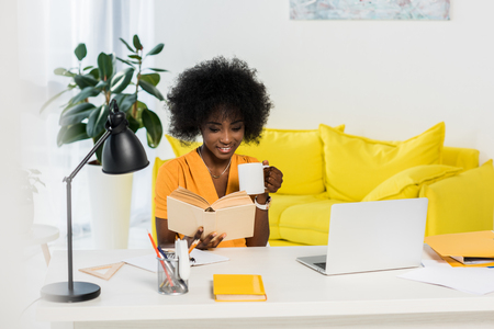 smiling african american woman with cup of coffee reading book at workplace with laptop at homeの写真素材