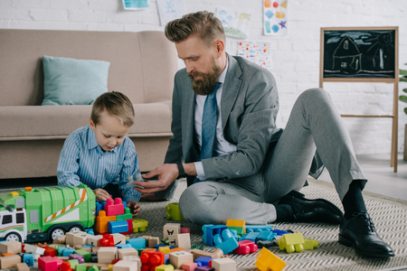businessman in suit and little son playing with colorful blocks together on floor at home, work and life balance conceptの写真素材