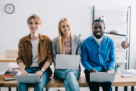 smiling multicultural businesspeople using laptops and sitting on table in modern office
