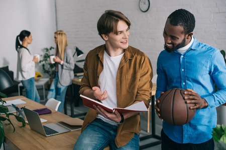 african american businessman holding ball and talking to partner with textbook and two businesswomen standing behind with coffeeの写真素材