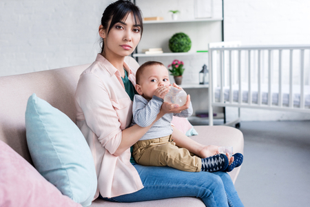 young beautiful mother feeding her little child with bottle at home and looking at cameraの写真素材