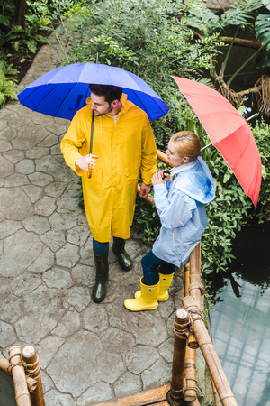 high angle view of young couple in raincoats with umbrellas spending time at park with exotic plantsの写真素材