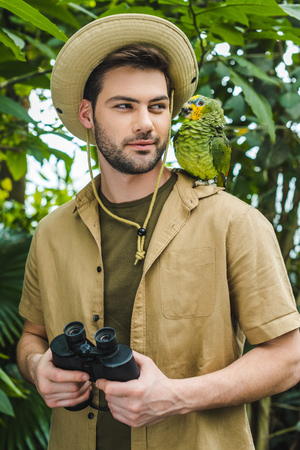 handsome young man in safari suit looking at parrot on shoulder in jungleの写真素材