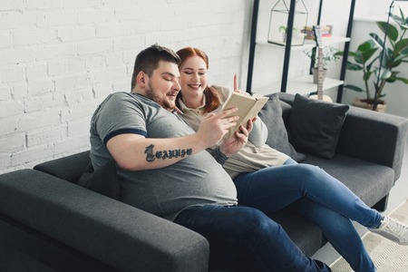 Smiling overweight boyfriend and girlfriend reading book on sofa at homeの写真素材