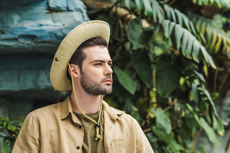 handsome young man in safari suit and hat looking away in jungleの写真素材