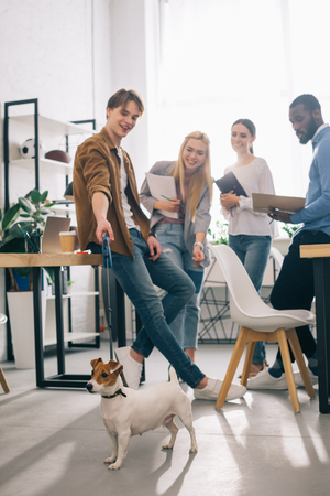 smiling multiethnic business colleagues with textbooks and jack russel terrier on leash in modern officeの写真素材