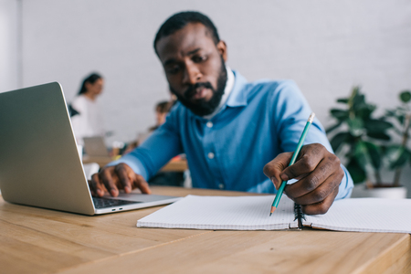 african american businessman writing in textbook and using laptop at table and colleagues standing behindの写真素材