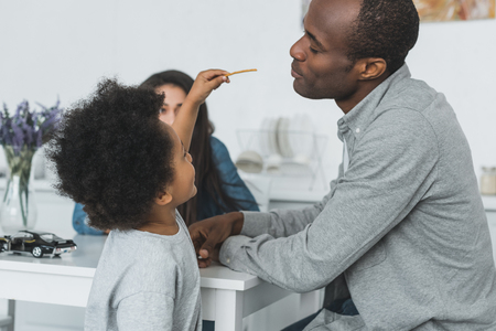 african american son feeding father with crispy breadsticks at homeの写真素材